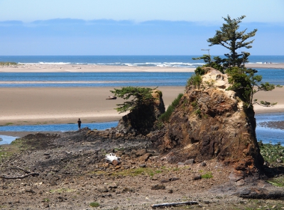 [Rocky foreground has one large rock formation and in the distance are bandss of water and sand and water and sand (due to the bay formation)before reaching the surf of the ocean.]